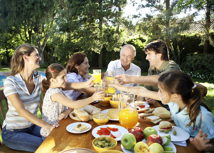 Family, including children (7-11) sitting at table in garden, toasting with orange juice and smiling Photograph by Maria Teijeiro