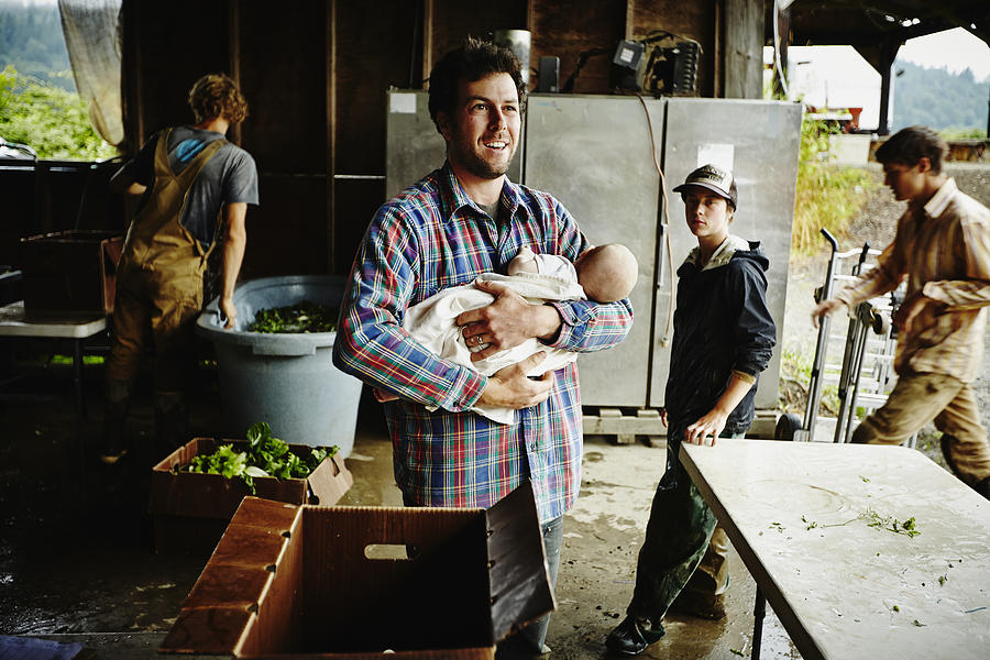 Farmer holding infant standing in work shed Photograph by Thomas Barwick