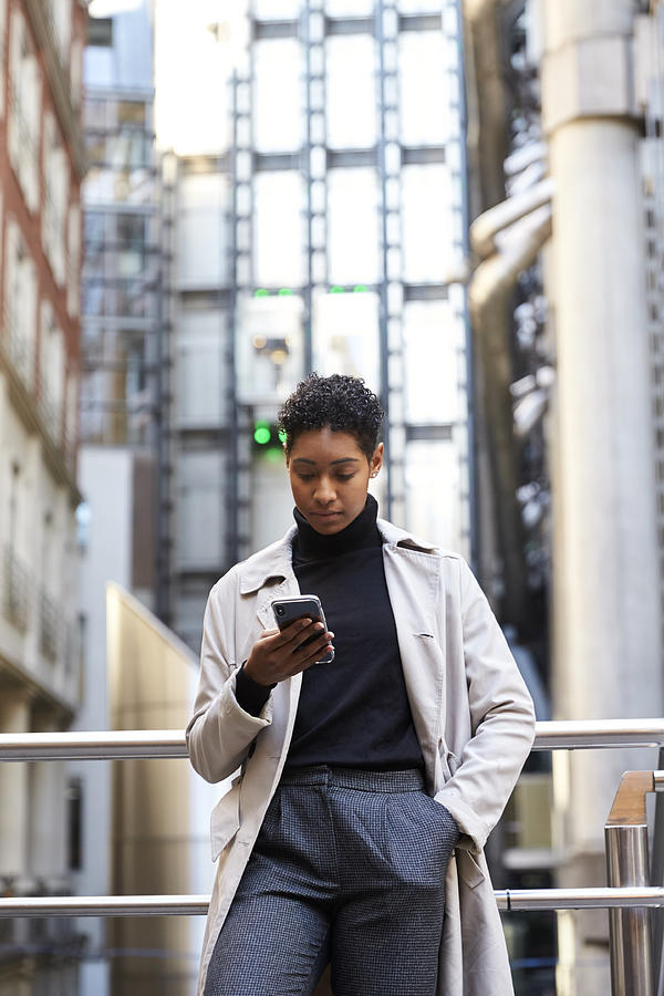 Fashionable young black woman standing in the city leaning on a hand rail using her smartphone, vertical Photograph by Monkeybusinessimages
