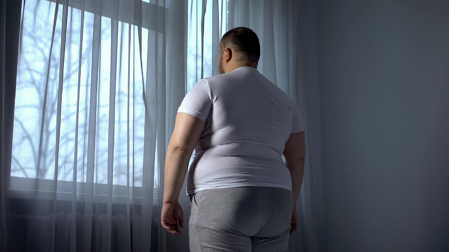 Fat man looking out through window, depressed introvert ashamed of obese body Photograph by Motortion