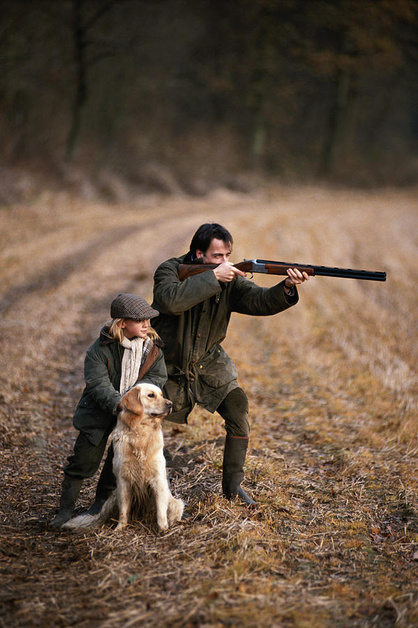 Father and daughter (4-7) in forest, man aiming with shotgun Photograph by David De Lossy