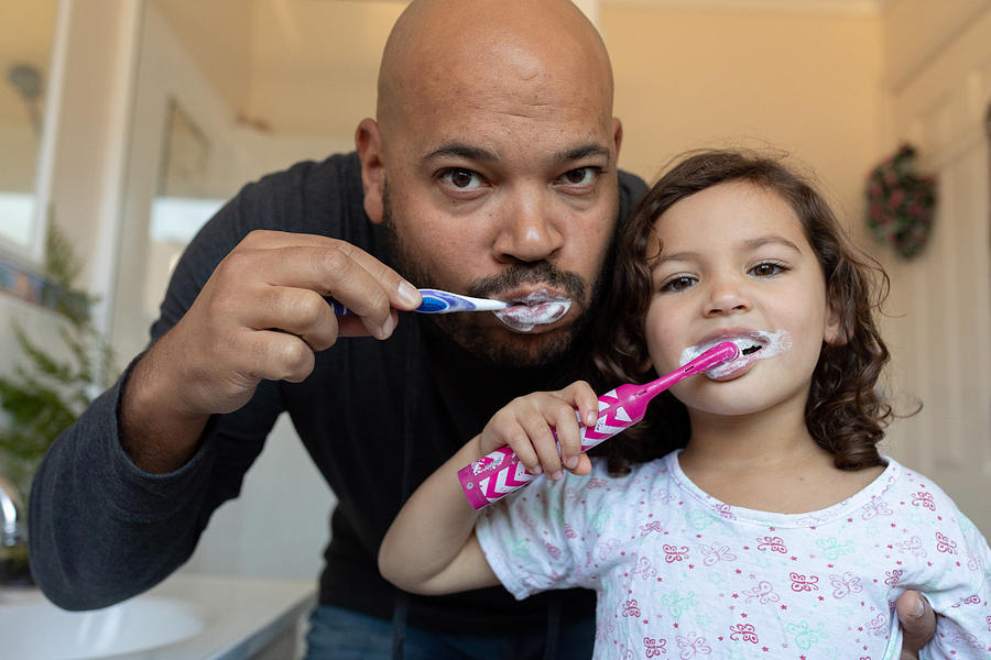 Father and daughter brushing their teeth and looking into camera Photograph by Jessie Casson