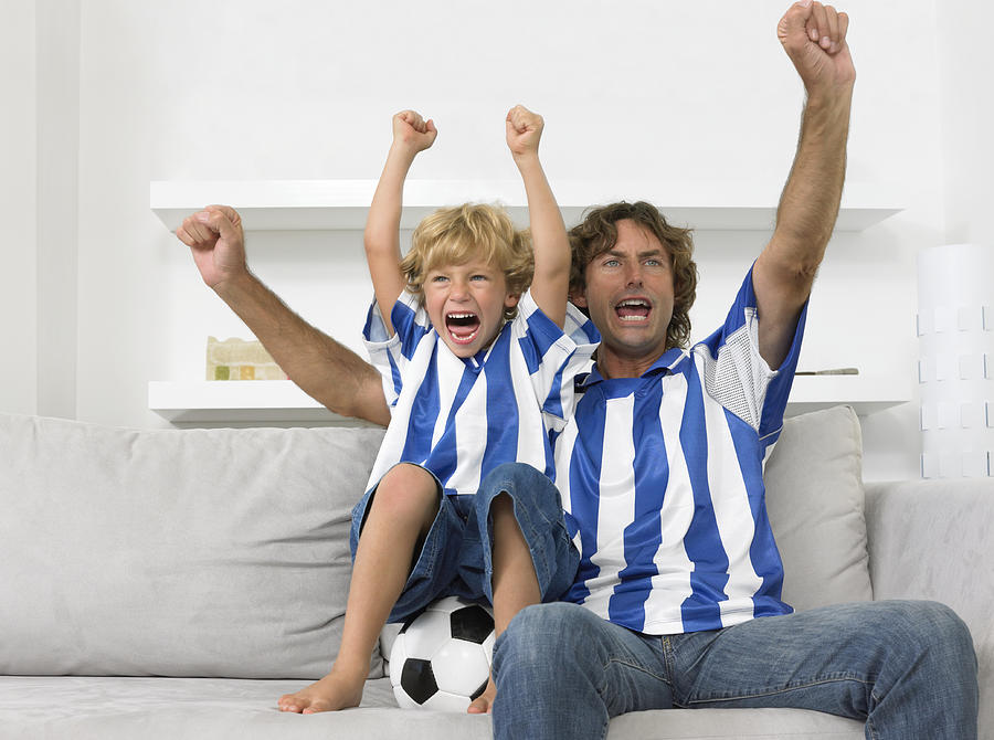 Father and Son Sitting on a Sofa, Wearing Football Strips and Cheering Photograph by Digital Vision.