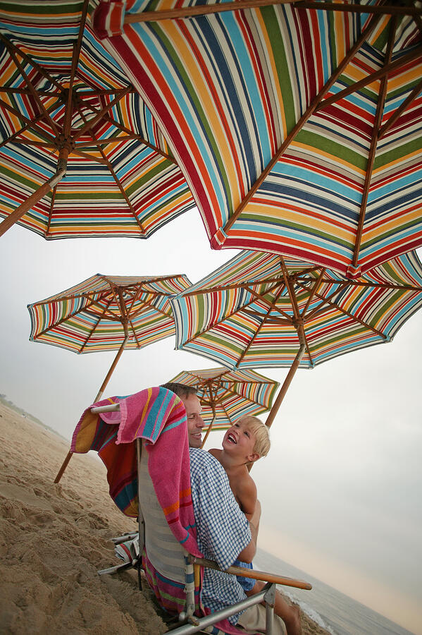 Father and son sitting together under striped beach umbrellas Photograph by Comstock