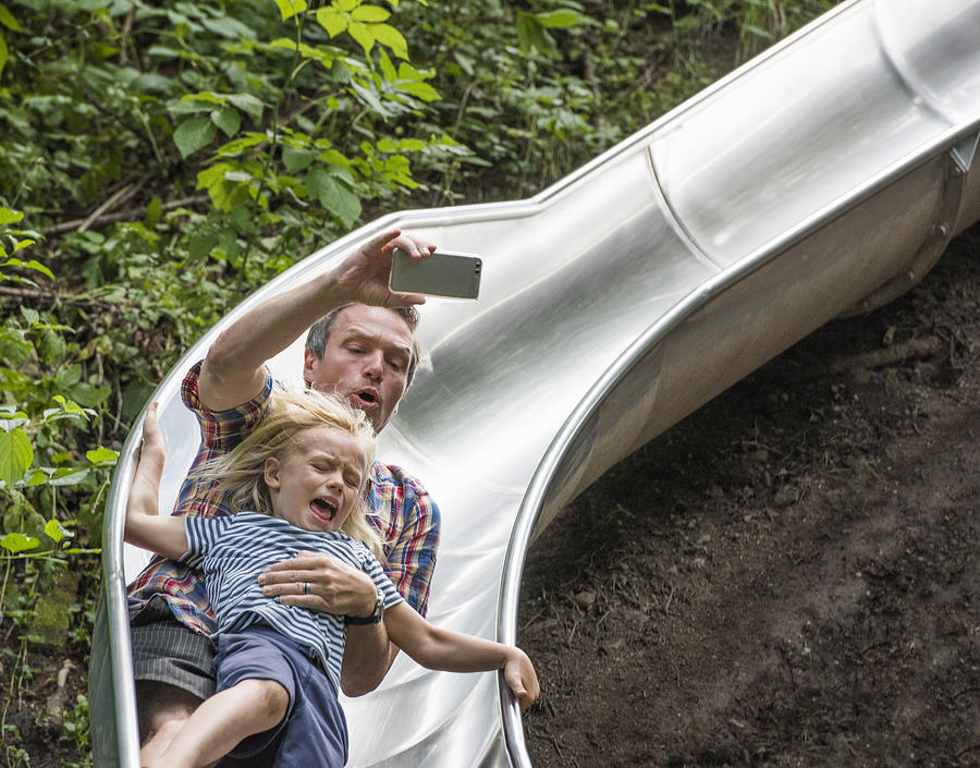 Father and son sliding down slide holding smartphone Photograph by Jlph
