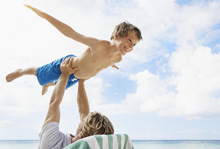 Father holding his son (10-12) mid-air while relaxing on deckchair on beach Photograph by Felix Wirth