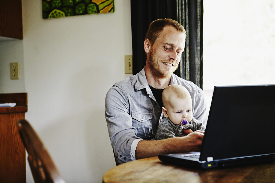 Father sitting working on laptop with baby on lap Photograph by Thomas Barwick