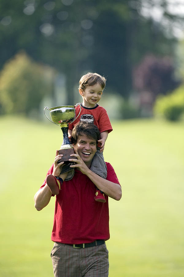 Father with son (2-3 years) on shoulders, holding trophy on golf course Photograph by Photo and Co