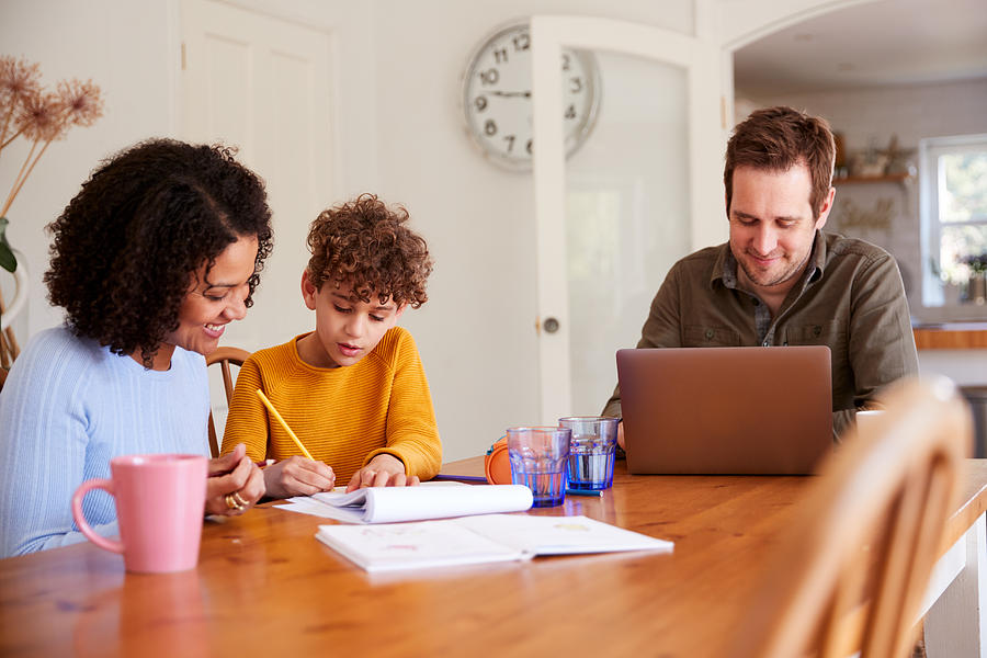 Father Works On Laptop As Mother Helps Son With Homework On Kitchen Table Photograph by Monkeybusinessimages