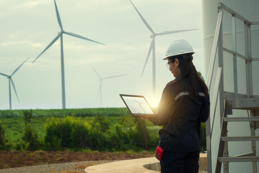 Female engineer wearing hard hat standing with digital tablet against wind turbine. Photograph by Tunvarat Pruksachat