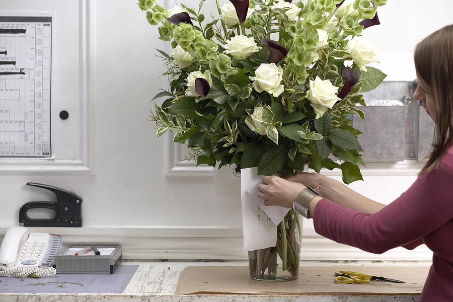 Female florist securing card to bouquet of flowers in vase, side view Photograph by David Leahy