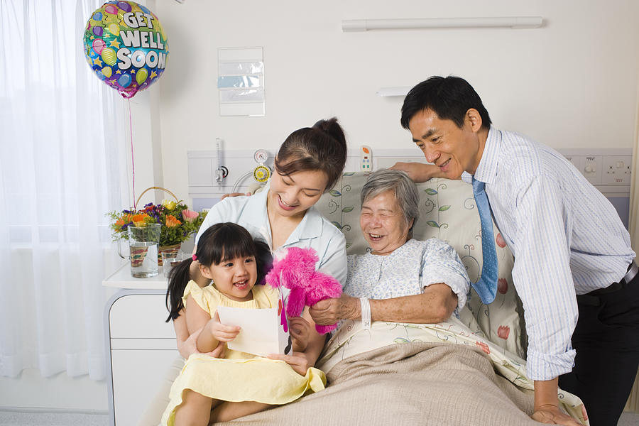 Female patient reclining on the bed and smiling with her family beside her Photograph by Asiaselects