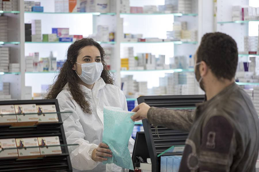 Female pharmacist wearing a surgical mask gives medication to the patient Photograph by Cekimdeyim