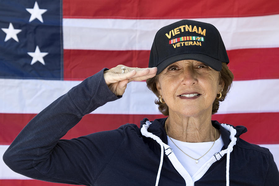 Female Vietnam Veteran Saluting  looking content wearing Veterans cap, with American Flag in background. Photograph by Johnrob