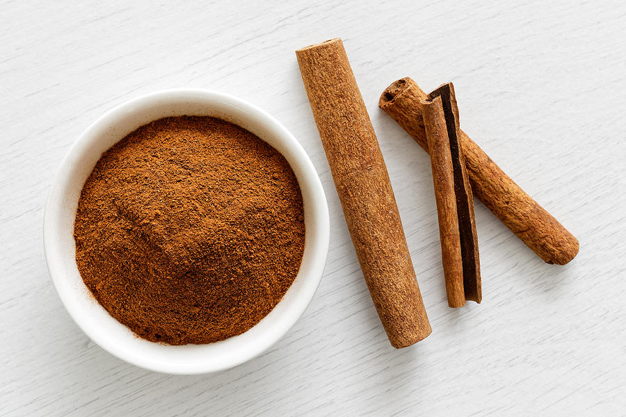 Finely ground cinnamon in white ceramic bowl isolated on white wood background from above. Cinnamon sticks. Photograph by Etienne Voss