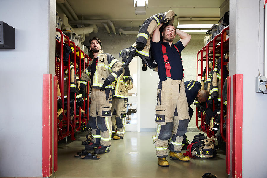 Firefighters wearing protective workwear in locker room while looking up at fire station Photograph by Maskot