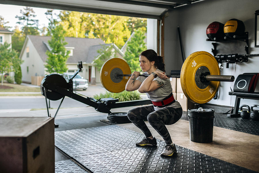 Fit woman performing front squat with heavy barbell in her home garage gym during covid-19 pandemic. Photograph by IngredientsPhoto