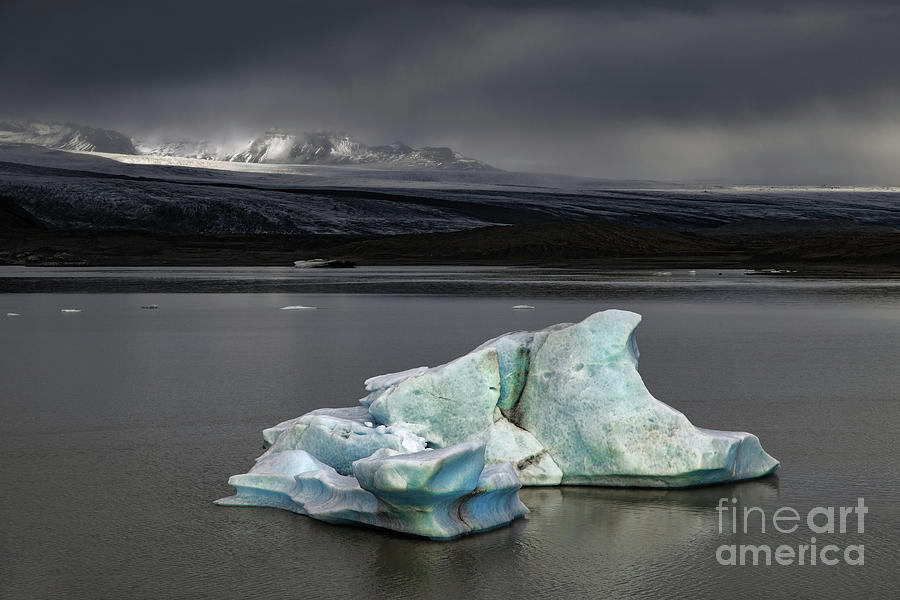 Fjallsarlon Glacier Lagoon, Iceland, on a stormy day. A blue iceberg floats in the lagoon and sunlight highlights the mountains behind. Photograph by Jane Rix