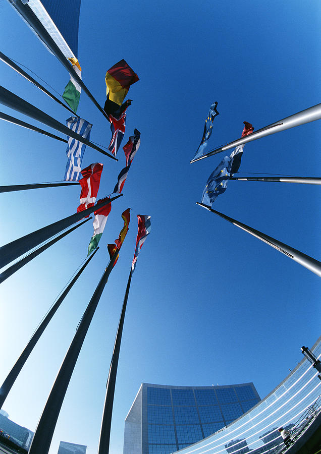 Flagpoles with flags from different countries, buildings in background, low angle shot Photograph by Christian Zachariasen