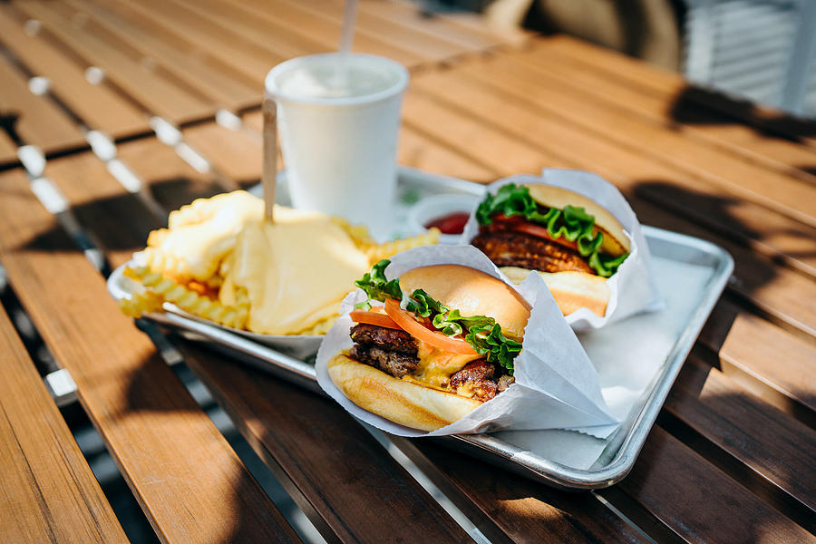 Freshly made beef burgers with cheese fries and soft drink on a tray served on a table with sunlight in an outdoor fast food restaurant Photograph by D3sign