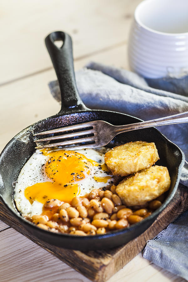 Fried egg, baked beans and hash browns in frying pan on wooden board Photograph by Westend61