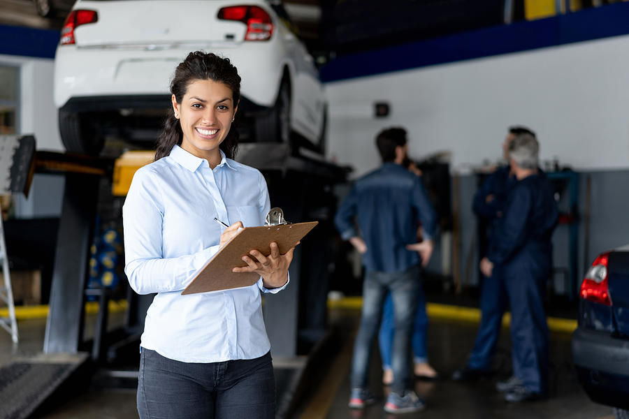 Friendly sales assistant at a car workshop holding a list on clipboard smiling at camera Photograph by Hispanolistic