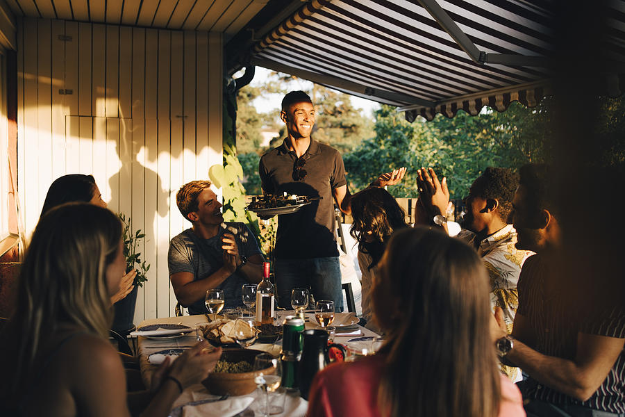 Friends at dining table clapping for man serving food in dinner party Photograph by Maskot