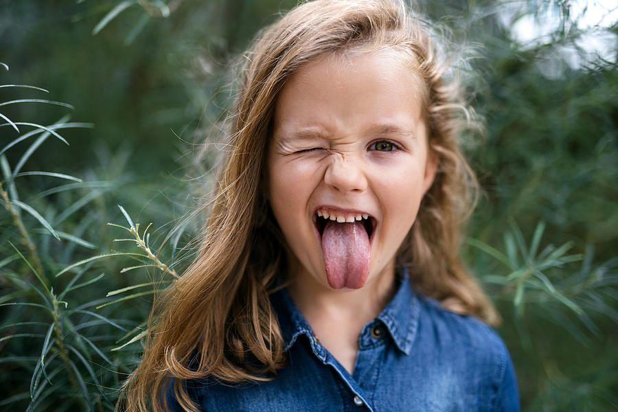 Front view portrait of small girl standing outdoors, sticking out tongue. Photograph by Halfpoint Images