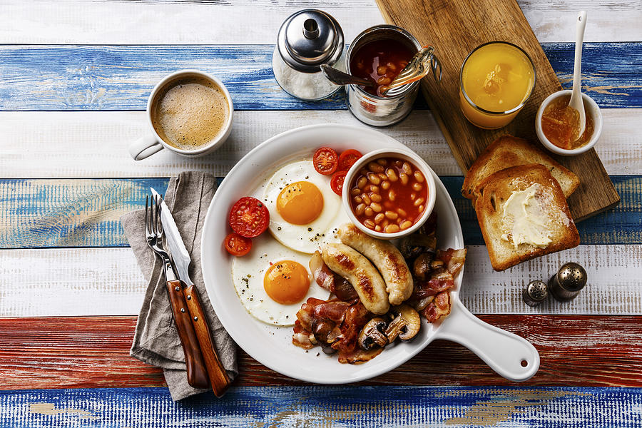 Full English breakfast in white pan with fried eggs, sausages, bacon, beans, toasts, orange fresh and coffee on wooden background Photograph by The Picture Pantry