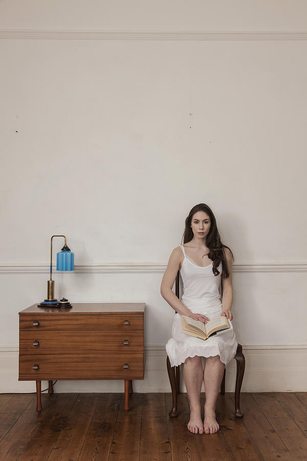 Full length front view of young woman sitting in chair wearing nightdress holding book looking at camera Photograph by Mark John