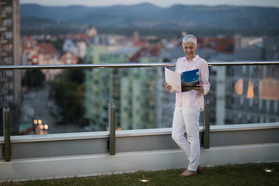 Full length of a smiling mature woman reading a magazine on a penthouse balcony. Photograph by Skynesher