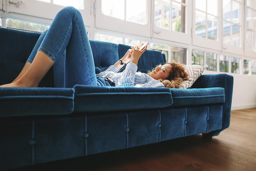 Full length of young woman using smart phone while lying on sofa at home Photograph by Cavan Images