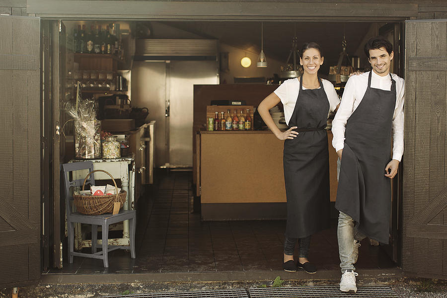 Full length portrait of smiling owners standing outside restaurant Photograph by Maskot