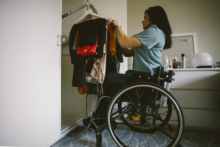 Full length side view of young disabled woman looking at clothes hanging in wardrobe Photograph by Maskot