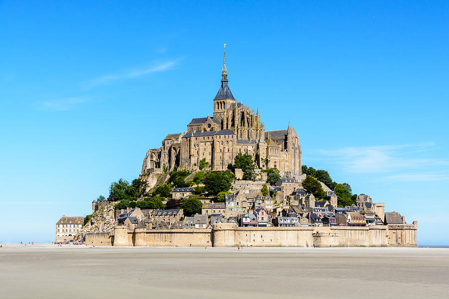 General view of the Mont Saint-Michel tidal island, located in France on the limit between Normandy and Brittany, with the exposed sand of the bay at low tide in the foreground under a blue sky. Photograph by Olrat