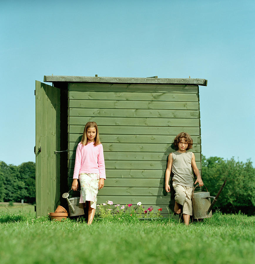 Girl (10-12) and boy (7-9) with watering cans leaning against shed Photograph by Colin Hawkins