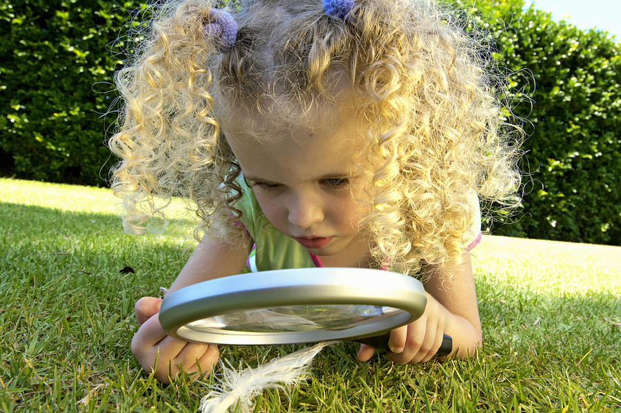 Girl (3-5) lying on grass, looking at feather through magnifying glass Photograph by Kane Skennar