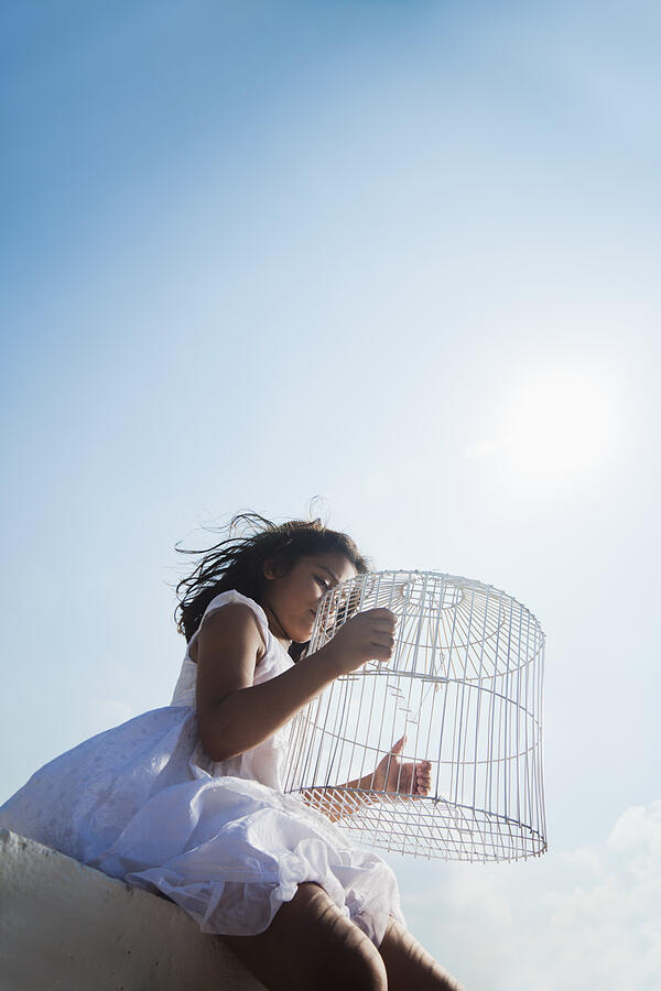 Girl (6-7) sitting on wall with empty birdcage, low angle view Photograph by ImagesBazaar