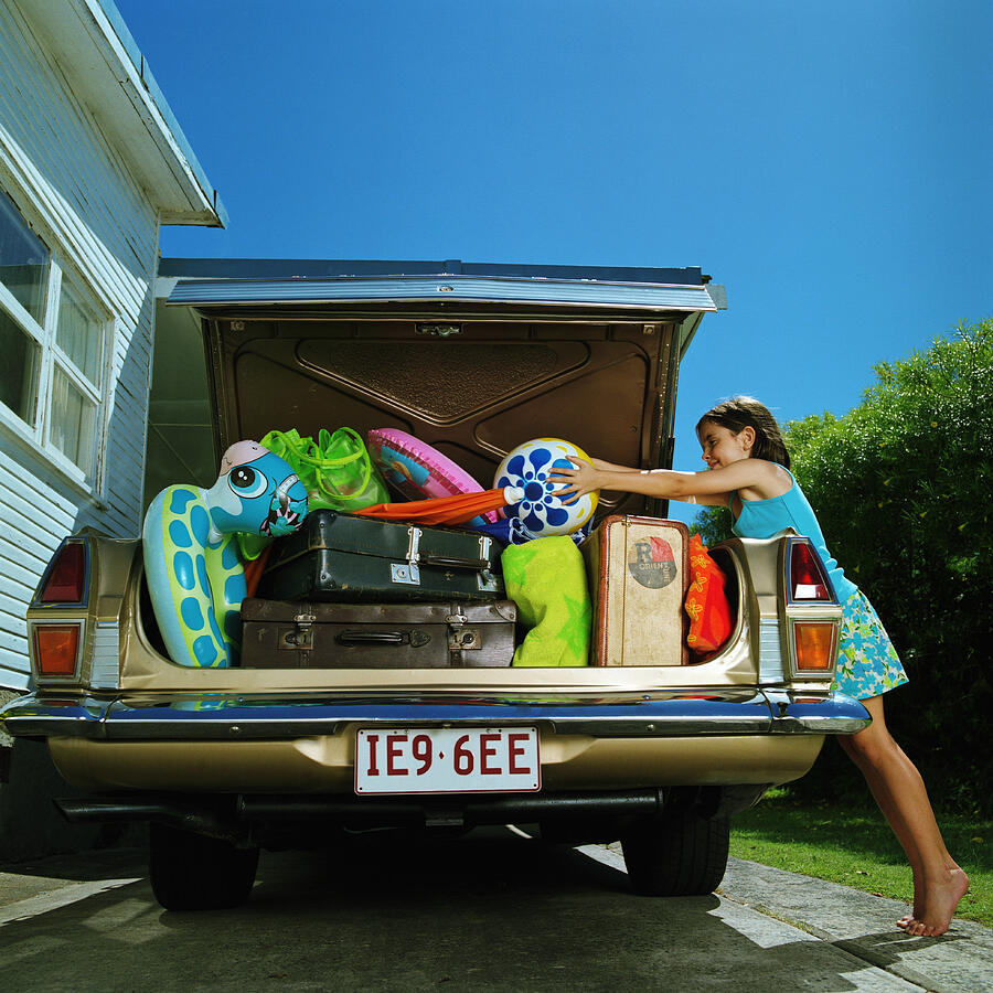 Girl (6-8) placing ball in heavilly loaded car boot Photograph by Peter Mason
