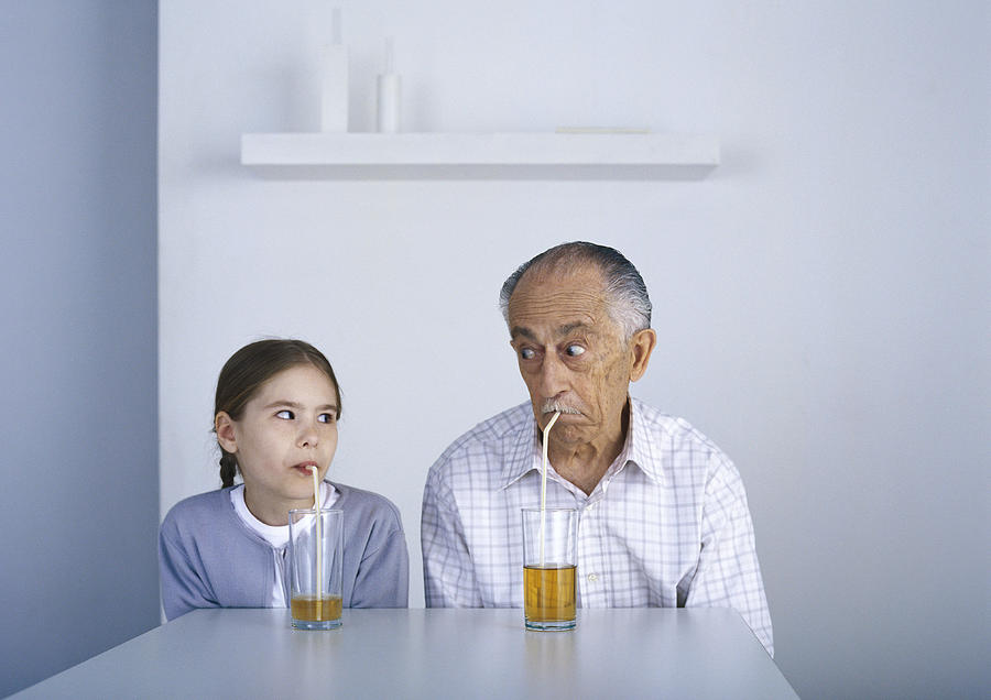 Girl and grandfather sitting at table drinking with straws Photograph by Laurence Mouton