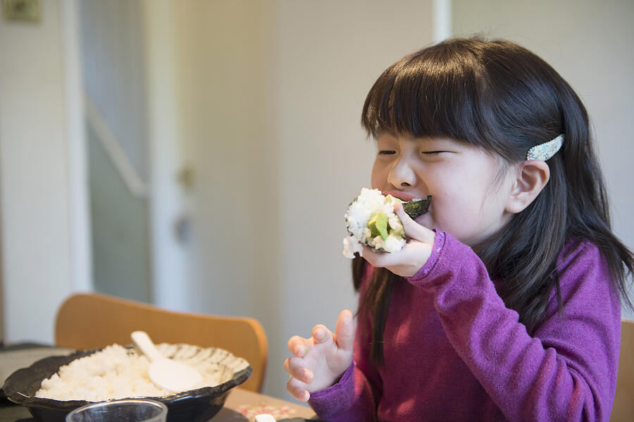 Girl eating large rice parcel at dining table Photograph by Kaori Ando