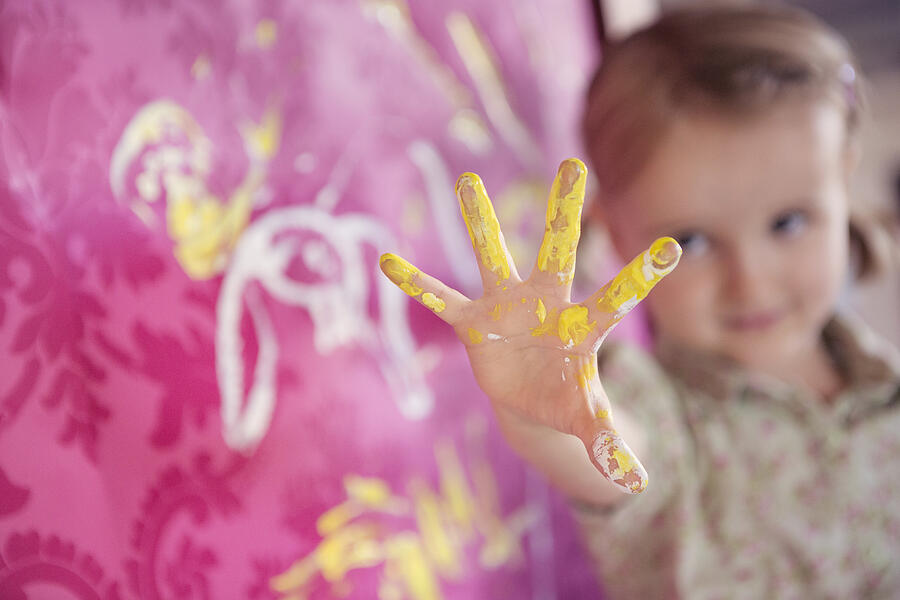 Girl holding up hand covered in finger paints, next to pink wallpaper Photograph by Elva Etienne