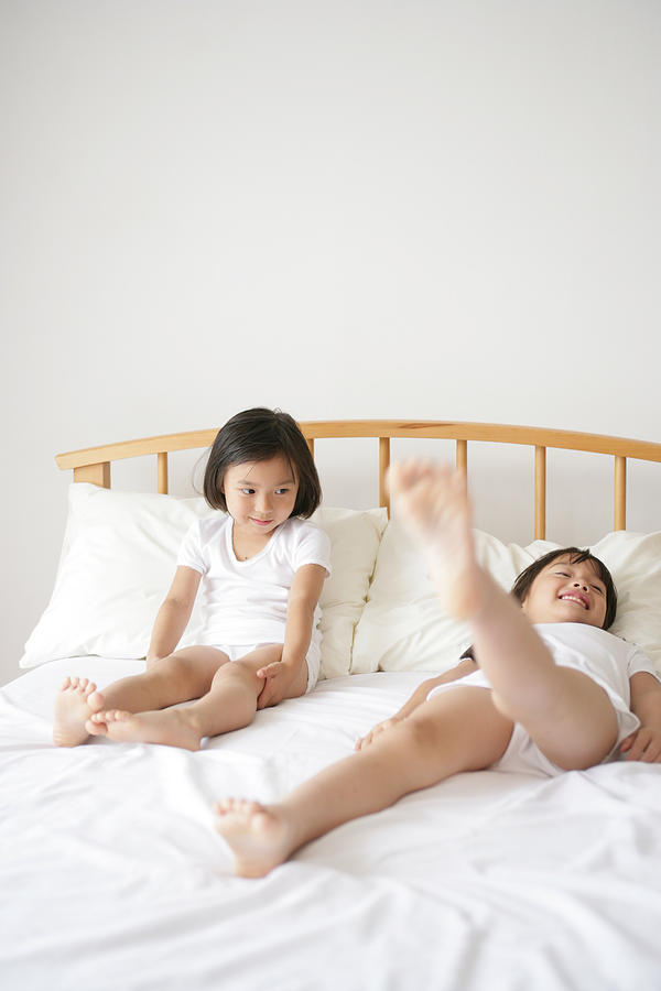 Girl lying on her back with one leg raised and her sister sitting beside her on the bed Photograph by Dex Image