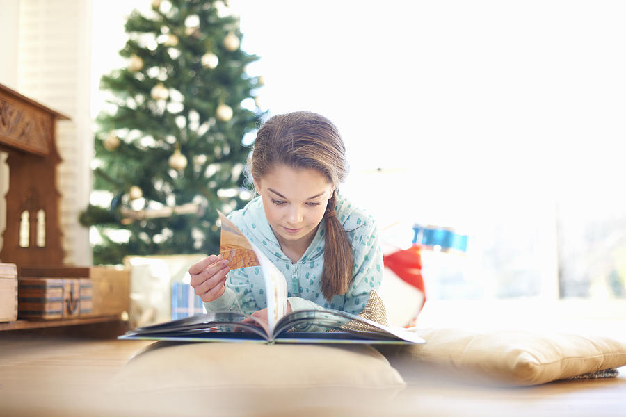 Girl lying on living room floor reading book at christmas Photograph by Peter Muller