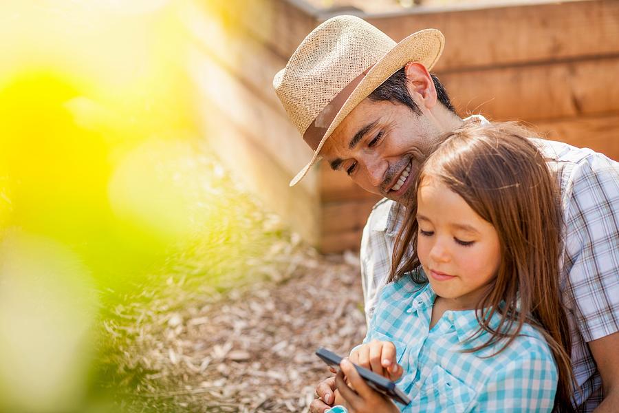 Girl on fathers lap using smartphone in community garden Photograph by Kevin Kozicki