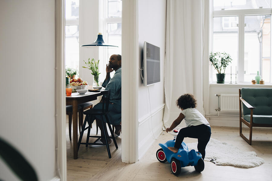 Girl riding toy car in living room while father having breakfast at dining table Photograph by Maskot