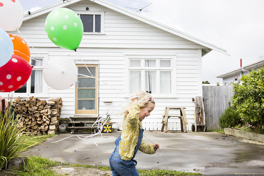 Girl runs past the front of a bungalow holding colourful balloons Photograph by Jessie Casson