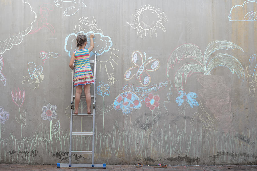 Girl standing on ladder drawing colourful pictures with chalk on a concrete wall Photograph by Westend61