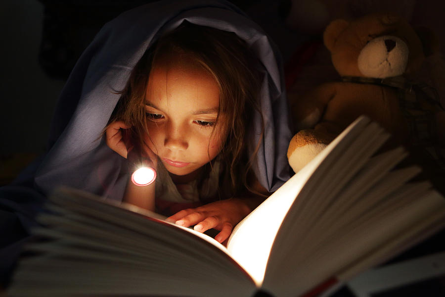 Girl under bed covers reading book by torchlight Photograph by Peter Cade