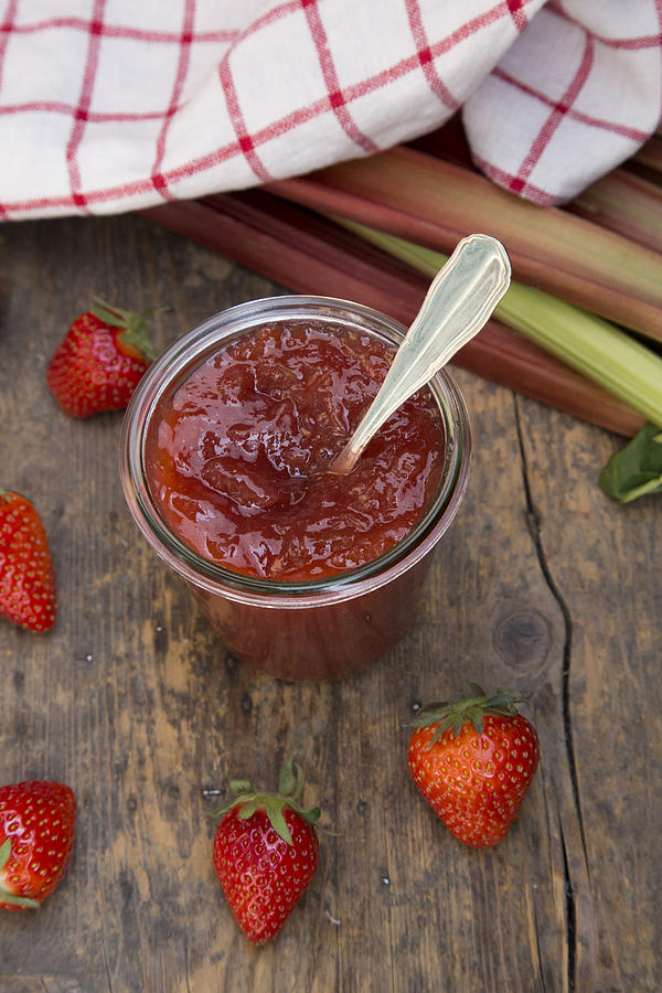 Glass of strawberry rhubarb jam, strawberries, kitchen towel and rhubarb on dark wood, elevated view Photograph by Westend61